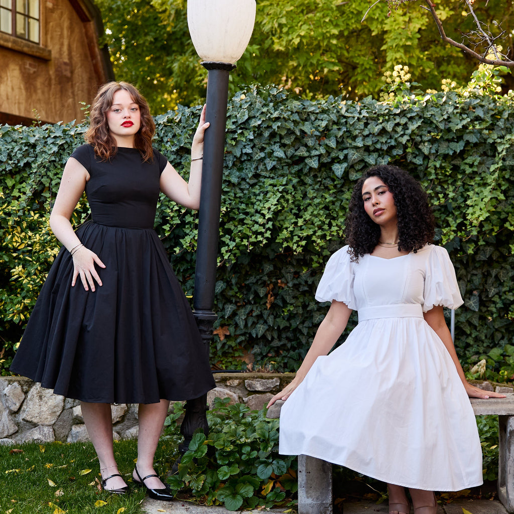 Two women in black and white dresses standing outdoors with a lamp post and stone bench.