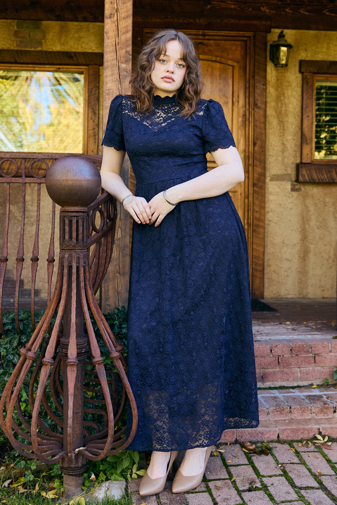 Woman in a navy blue dress standing on a porch with wooden beams and a lantern - navy lace midi elegant romantic bridal shower church wedding guest outfit modest
