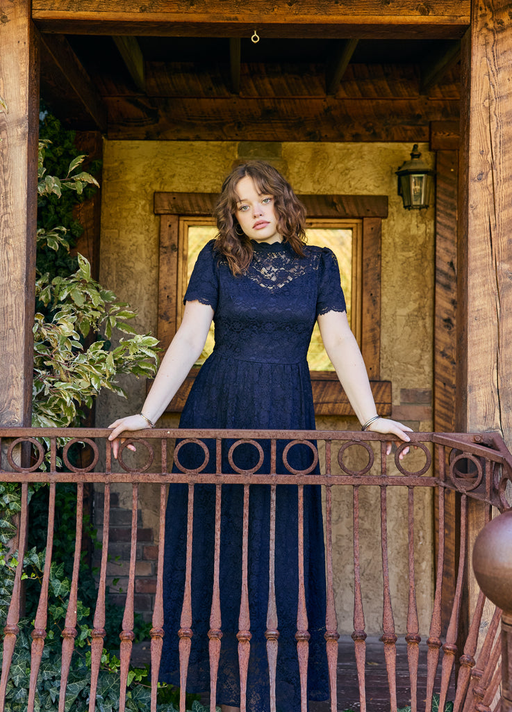 Woman in a navy blue dress standing on a balcony with wooden beams and greenery - navy lace midi elegant romantic bridal shower church wedding guest outfit modest