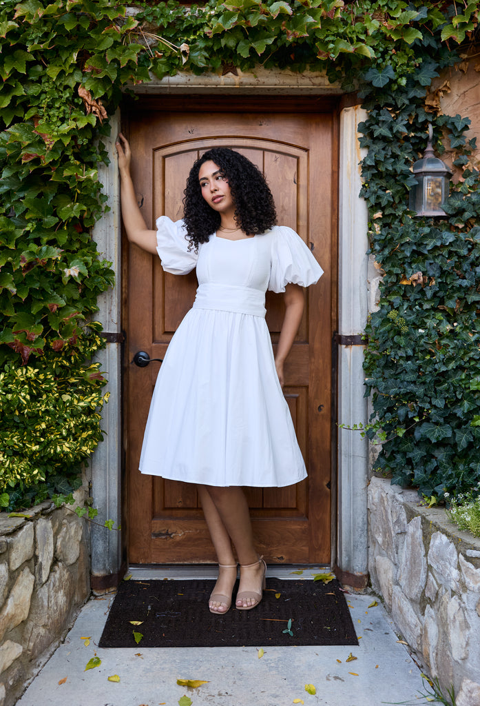 Woman in a white dress standing in front of a wooden door with greenery - cotton puff sleeve white