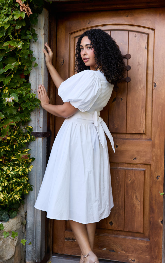 Woman in a white dress standing in front of a wooden door with greenery on the side - cotton puff sleeve white