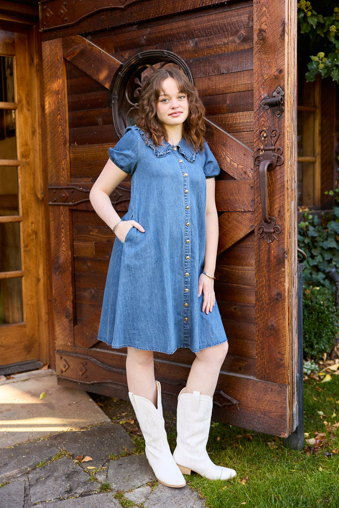 Young girl in a blue dress and white boots standing in front of a wooden door -blue denim midi button down collared casual summer everyday jean picnic outfit modest
