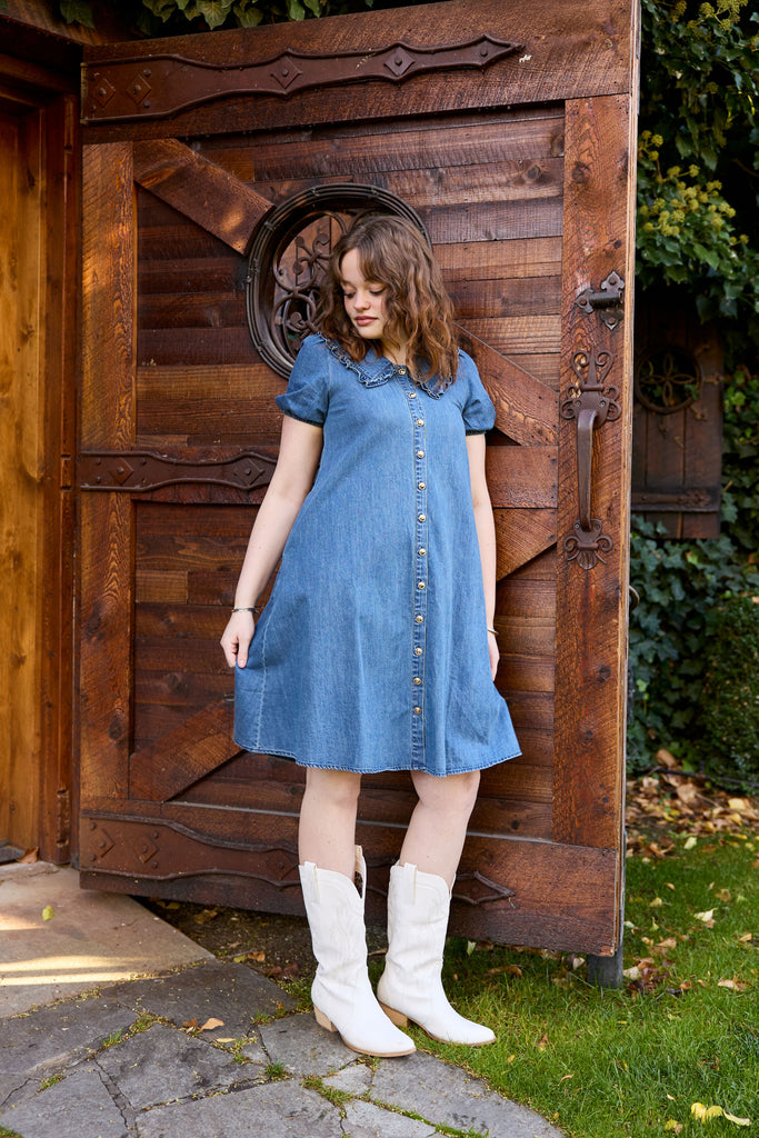 Woman in a blue dress and white boots standing in front of a wooden door -blue denim midi button down collared casual summer everyday jean picnic outfit modest