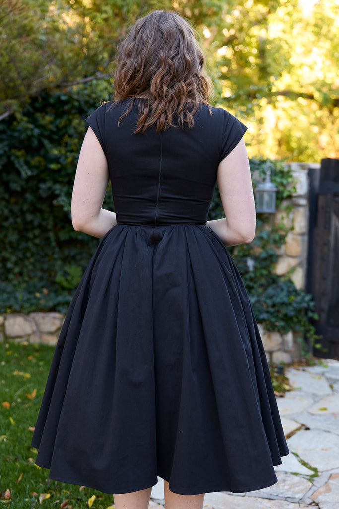 Woman wearing a black dress standing outdoors with greenery in the background - black 50s fit and flare vintage midi 1950s inspired timeless for events holiday party retro tea-length