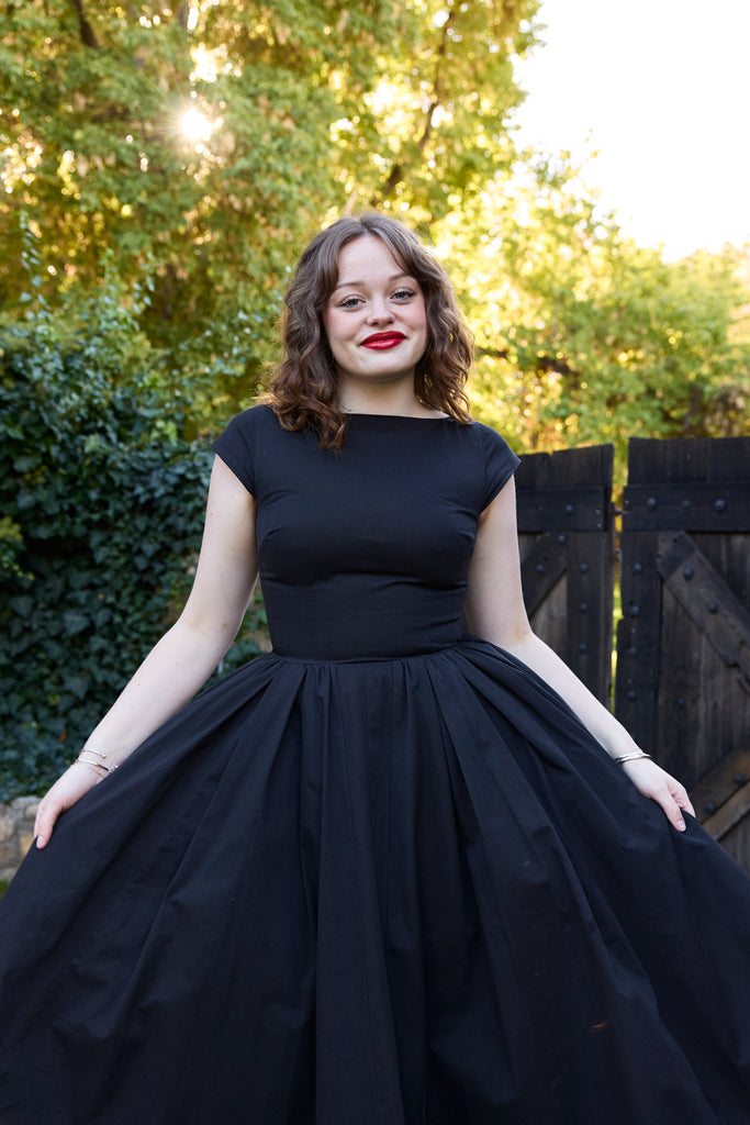 Woman wearing a black dress standing outdoors with greenery in the background - black 50s fit and flare vintage midi 1950s inspired timeless for events holiday party retro tea-length