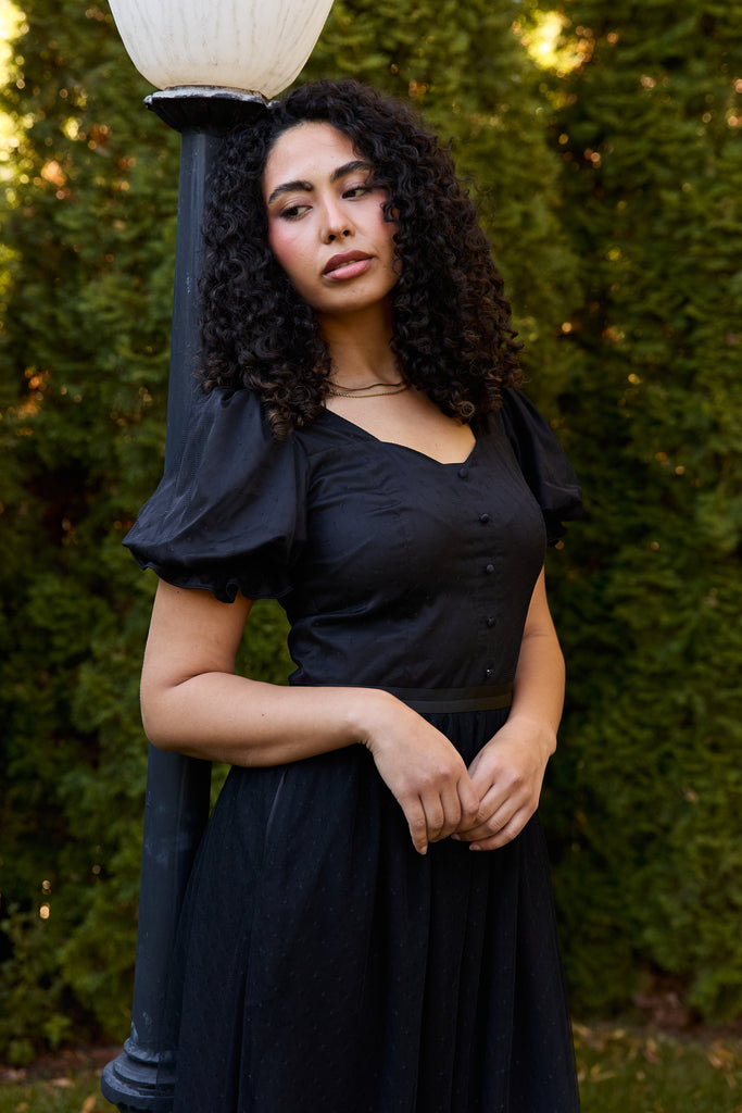Woman in a black dress standing outdoors with greenery in the background - black ballerina modest evening cocktail wedding guest