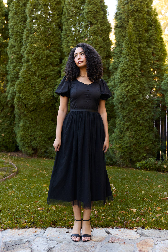 Woman in a black dress standing outdoors with green trees in the background - black ballerina modest evening cocktail wedding guest