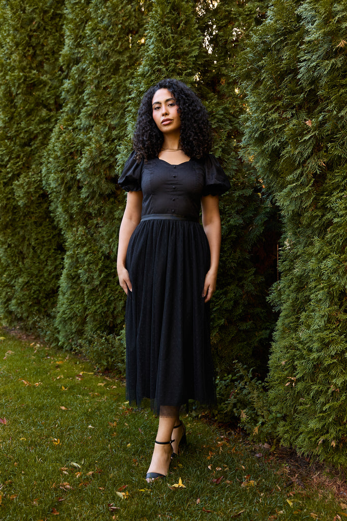 Woman in a black dress standing between two tall green bushes - black ballerina modest evening cocktail wedding guest